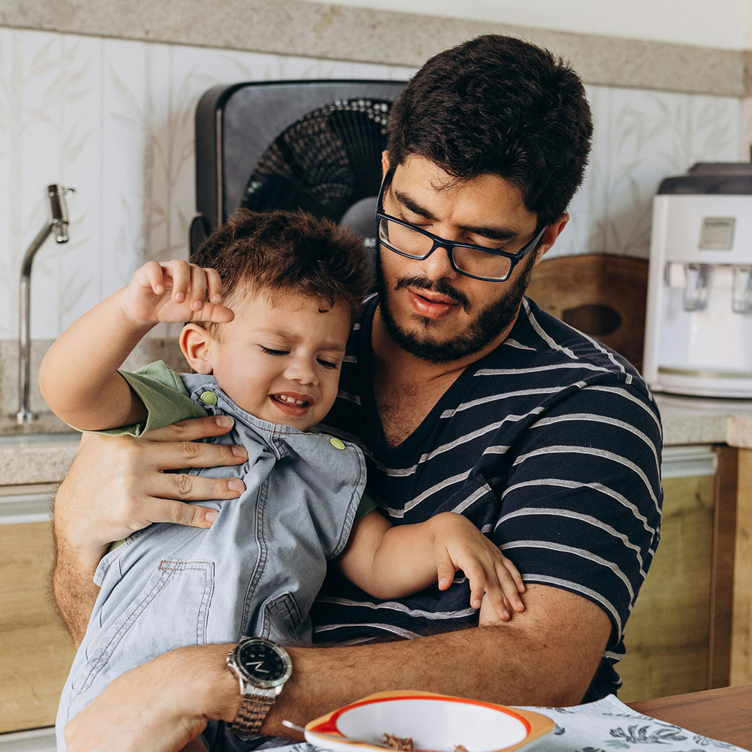 A father in glasses holds his young son in the kitchen during a quiet morning moment