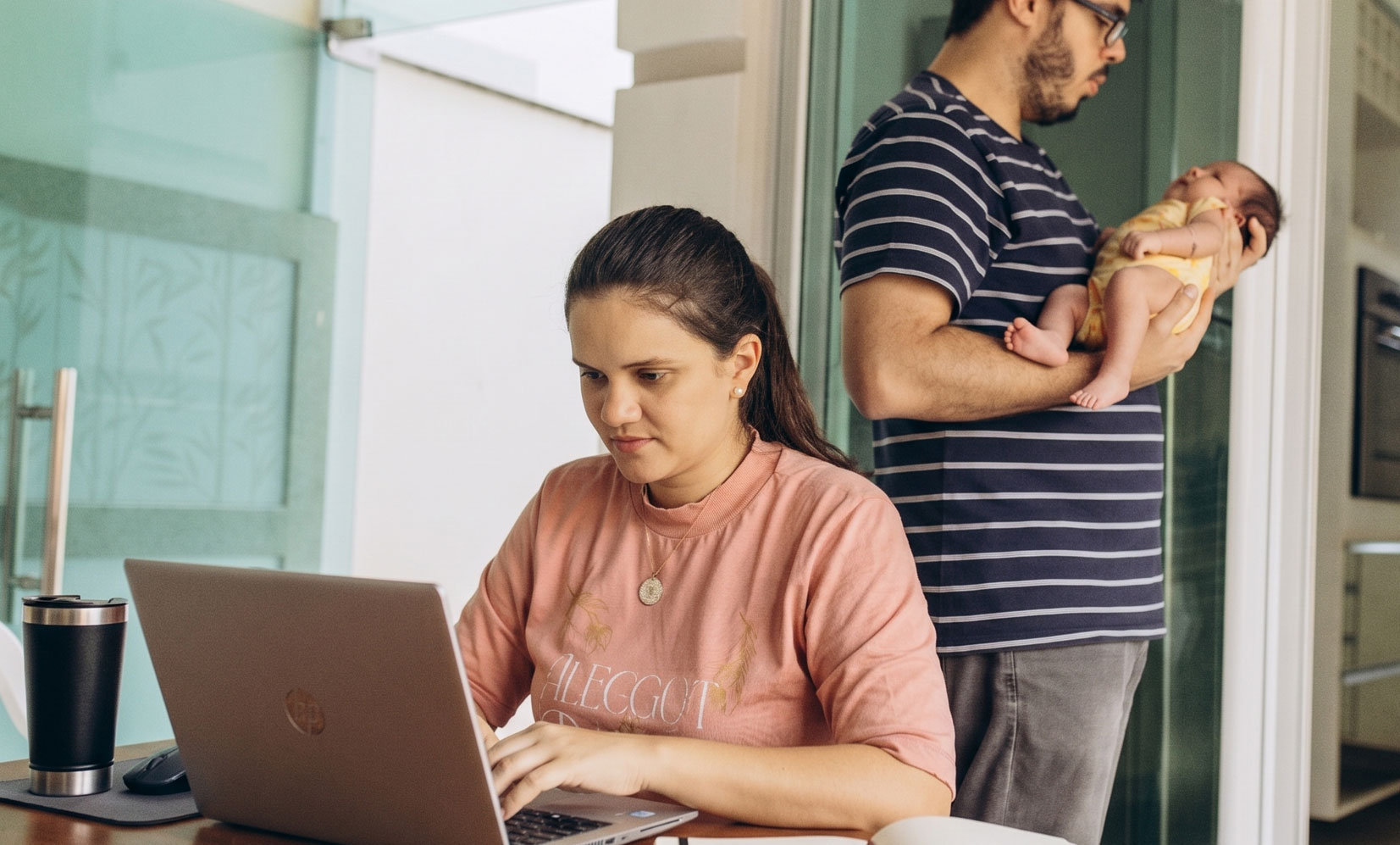 A mother works on her laptop at the kitchen table while a father holds their infant in the background