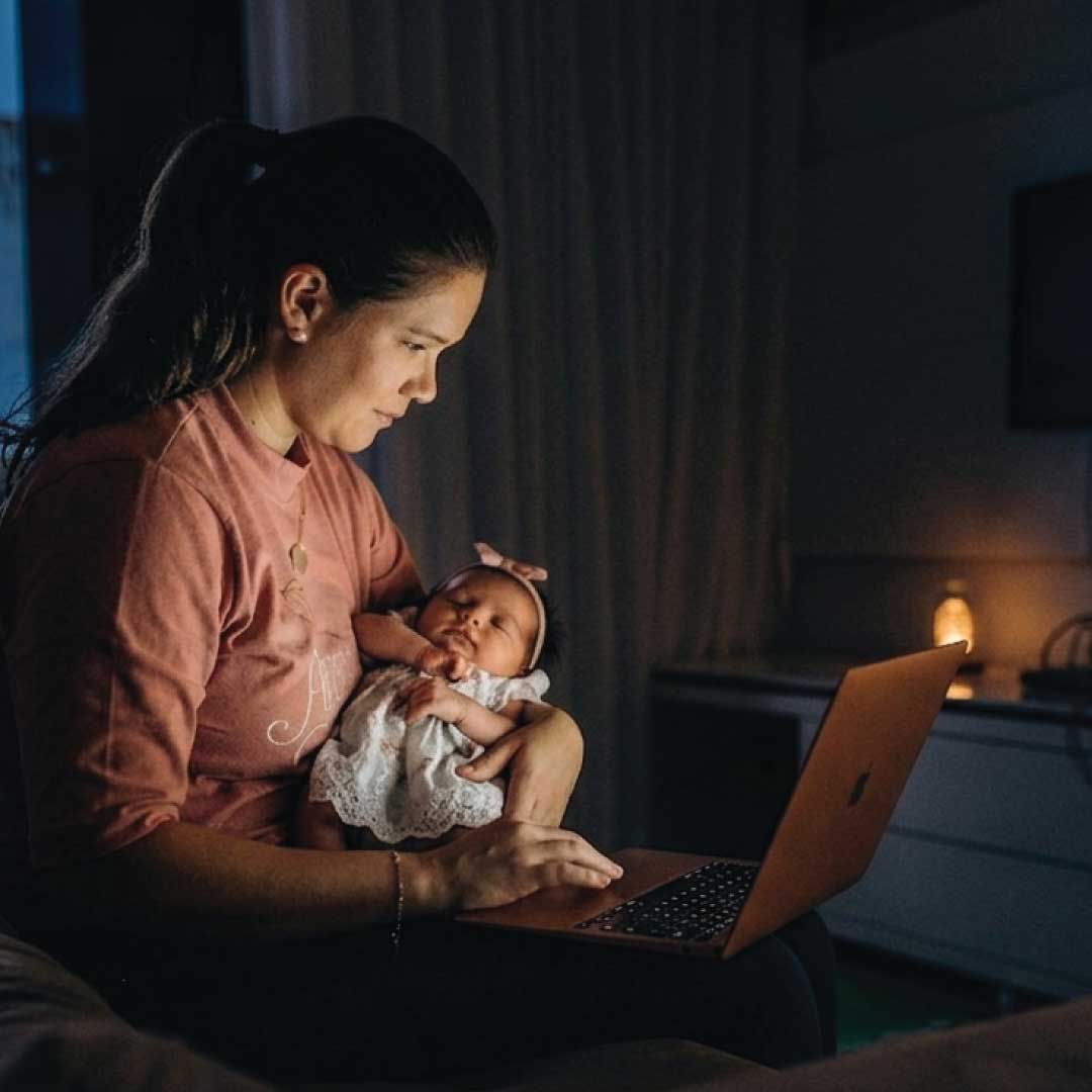 A mother holds her sleeping infant while working on a laptop late at night in a softly lit room