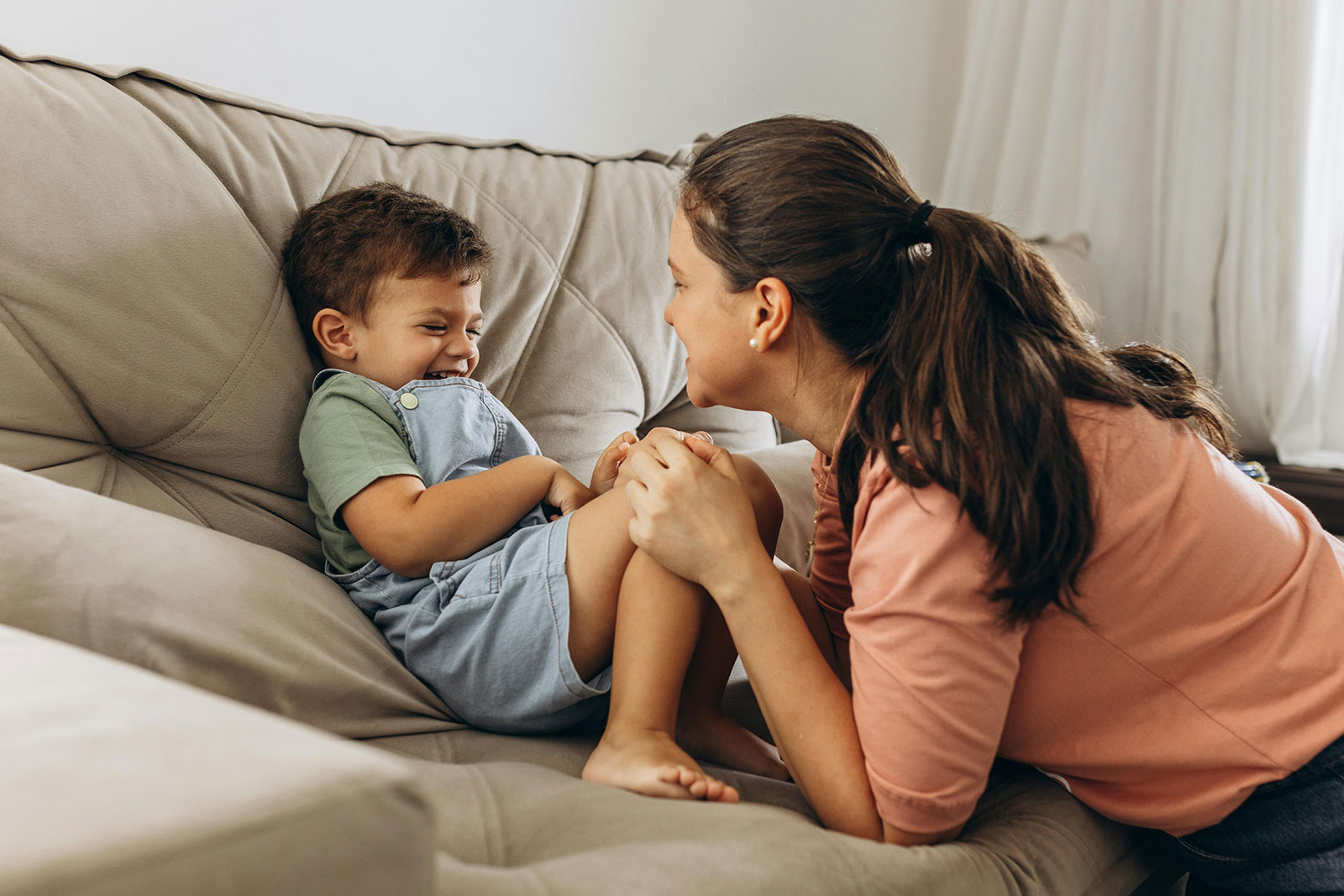 A mother leans in close, smiling and laughing with her young son on a couch